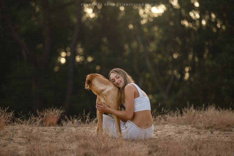 Hunde. Frau mit Hund. Der Hund ist beige und blickt in die Ferne. Die Frau hat die Augen geschlossen und lehnt sich an den Hund an.